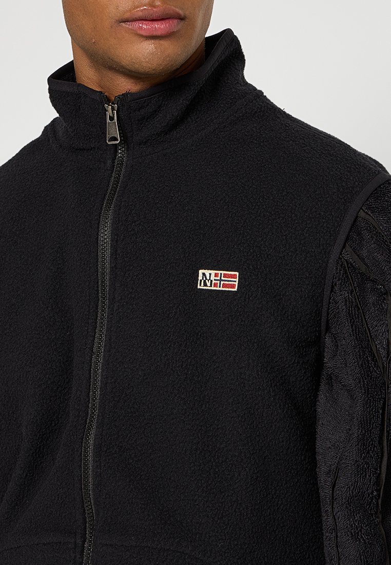 Veste en polaire noire avec un col haut et une fermeture éclair. Présente un patch avec des accents rouges et blancs sur la poitrine et des manches noires texturées.