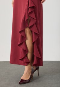 Bordeauxrotes Kleid mit rüschengeschnittenem Seitenschlitz, weichem Stoff, begleitet von spitzen Absatzschuhen in tiefem Burgunderrot und Grau mit glänzender Oberfläche.