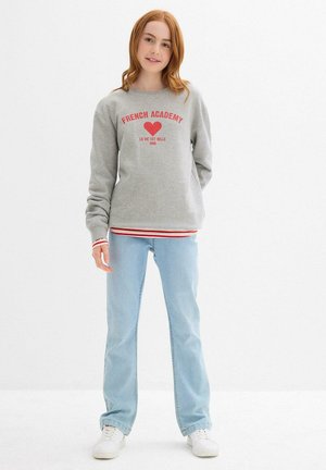 Junge Frau mit roten Haaren, die einen grauen Pullover mit rotem Text und Herz, hellblauen Jeans und weißen Sneakers trägt, steht vor einem weißen Hintergrund.