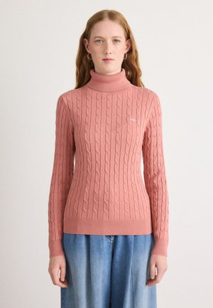 Pink cable-knit turtleneck sweater with ribbed hem and cuffs. Features a subtle logo detail on the left chest area. Worn with light blue denim.