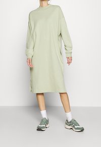Jasnozielona, oversize sukienka-sweterek z długimi rękawami, okrągłym dekoltem i bocznymi rozcięciami, noszona z zielonymi trampkami i białymi skarpetkami sięgającymi kostek.