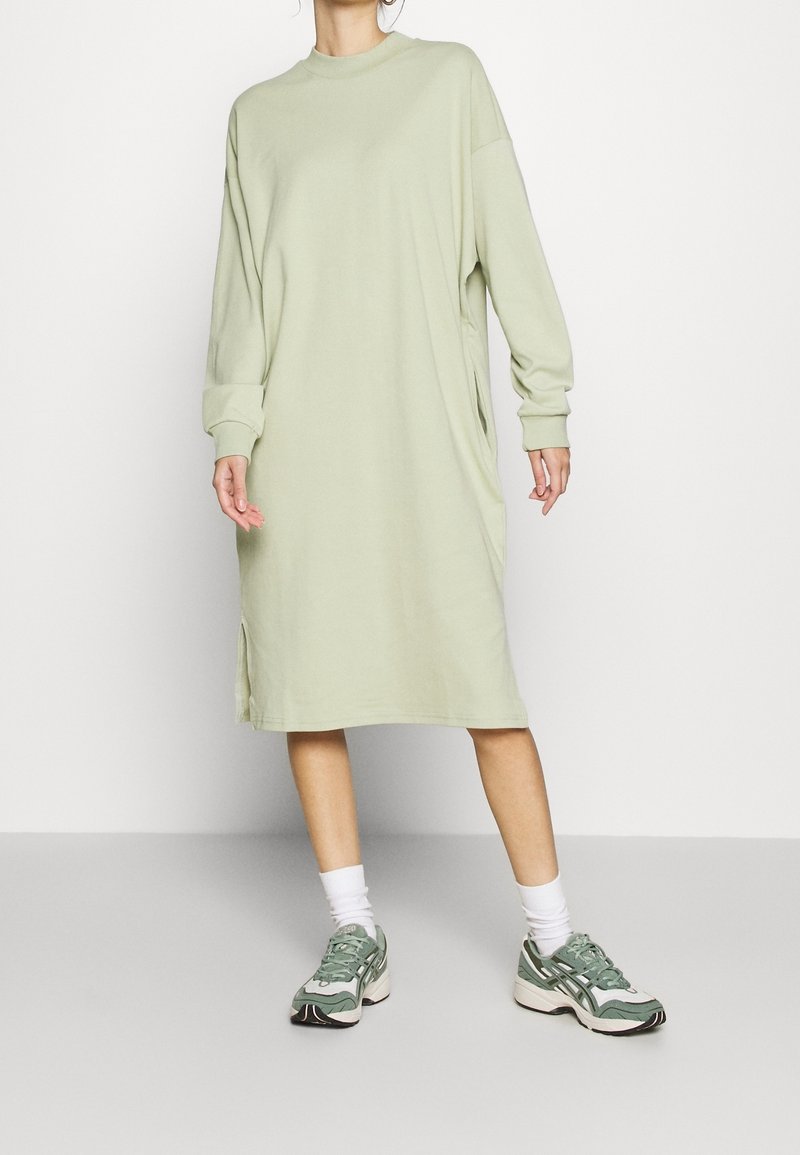 Jasnozielona, oversize sukienka-sweterek z długimi rękawami, okrągłym dekoltem i bocznymi rozcięciami, noszona z zielonymi trampkami i białymi skarpetkami sięgającymi kostek.