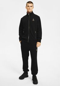 Mężczyzna ubrany w czarną kurtkę na zamek i dopasowane spodnie dresowe z logo na piersi i udzie, stojący z rękami częściowo w kieszeniach.