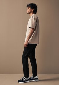 Beiges Baumwoll-T-Shirt mit kurzen Ärmeln, kombiniert mit eng anliegenden schwarzen Hosen und navyblauen Sneakers mit grauen Akzenten und weißem Logo.