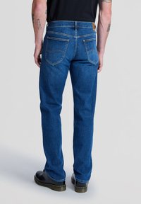 Mężczyzna ubrany w niebieskie jeansy o prostych nogawkach i czarne buty stoi tyłem do kamery na jednolitym szarym tle.