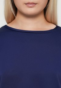 Žena s dugom plavom kosom koja nosi tamnoplavu majicu, prikazuje vrat, vilicu i usne bez vidljivog izraza na licu.