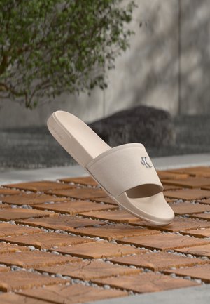 Mule beige Calvin Klein avec bride en tissu et logo, flottant au-dessus d'un sol extérieur en carreaux de bois et galets.