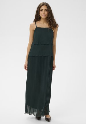 Woman with long curly hair wearing a dark green pleated sleeveless maxi dress and black pointed shoes, standing on plain background.