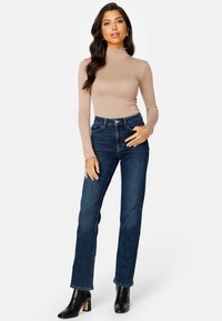 Krémový top s dlouhými rukávy a volánkovým límcem, spárovaný s jeansy s vysokým pasem v tmavě modré barvě a černými kotníkovými botami na podpatku. Hladká látka a přiléhavý střih.