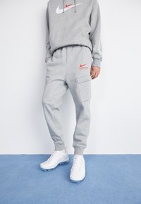 Šedá bavlněná mikina s bílým logem Nike a červeným textem "Athletic Dept", v kombinaci se šedými tepláky se zipovými kapsami a bílými sportovními botami.