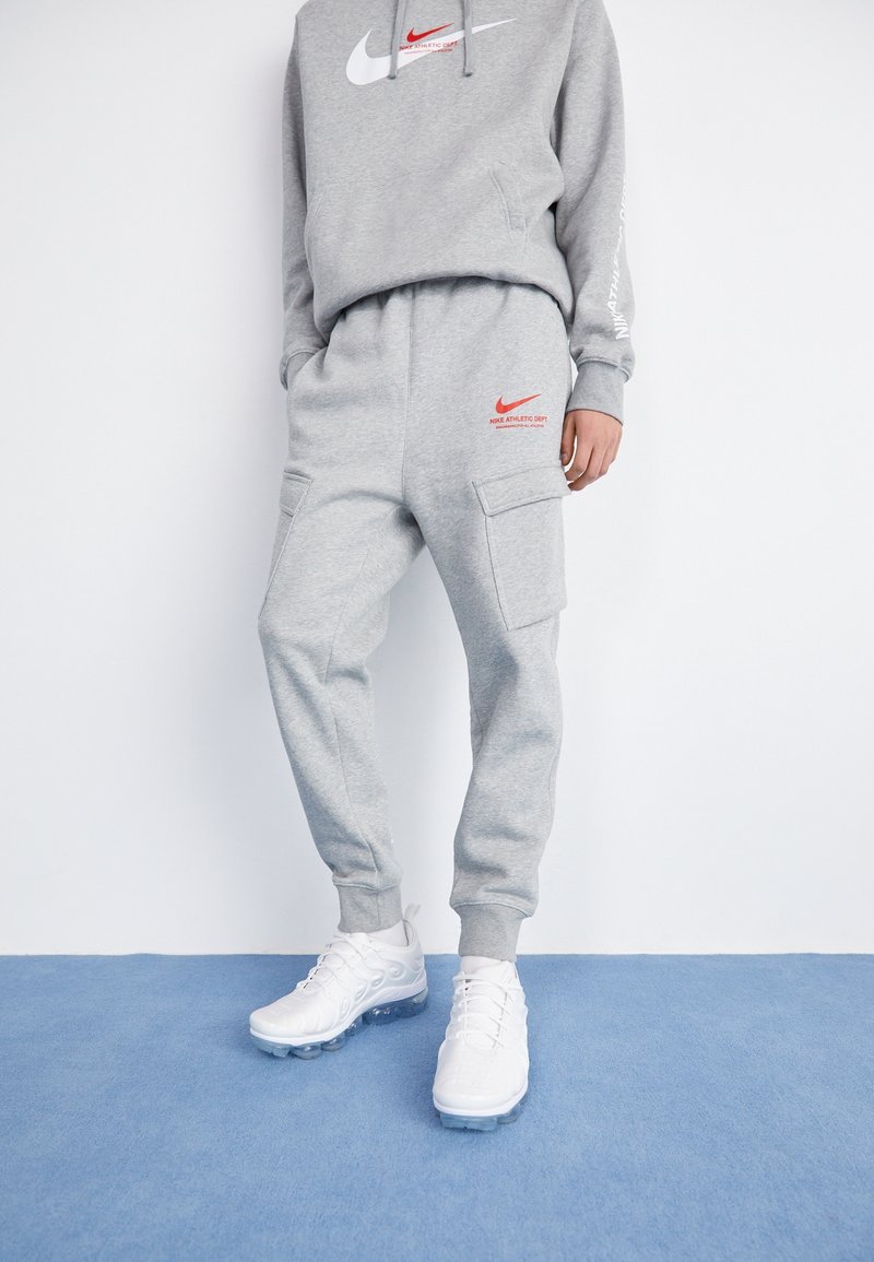Šedá bavlněná mikina s bílým logem Nike a červeným textem "Athletic Dept", v kombinaci se šedými tepláky se zipovými kapsami a bílými sportovními botami.
