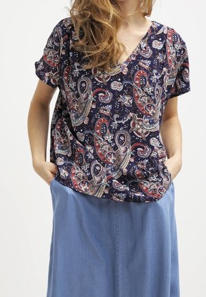 Vrouw draagt een marineblauw paisley blouse met korte mouwen, ingestopt in een lichtblauwe rok, met handen in de zakken.