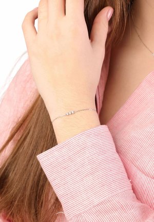 Bracelet en chaîne argentée avec trois accents ronds, porté sur un poignet, associé à une chemise à rayures roses. Texture métallique lisse, design délicat.