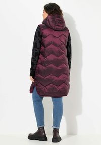 Gilet imbottito bordeaux con cappuccio, decorato con un motivo a zig-zag. Abbinato a jeans blu e stivaletti neri.