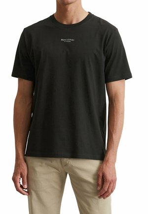 Mann trägt ein schwarzes Marc O’Polo T-Shirt mit beigen Hosen, steht mit entspannten Armen an den Seiten.