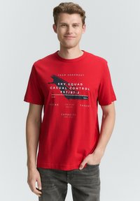 Camiseta de algodón roja con cuello redondo, mangas cortas y texto gráfico en blanco y negro. Combinada con jeans gris oscuro.