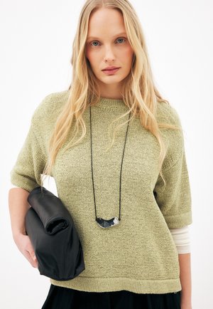 Lyshåret kvinde iført lysegrøn strikket sweater og lang sort halskæde, der holder en foldet sort læderclutch under venstre arm.