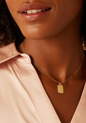 Gouden hanger ketting met een rechthoekig label aan een dunne ketting, gedragen boven een lichtroze satijnen shirt met zichtbare kraag. Gladde textuur.