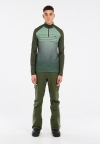 Groene blouse met lange mouwen en een ritskraag met een verloop-effect, gecombineerd met bijpassende groene broek en zwarte laarzen. Eenvoudig ontwerp, zonder patronen.