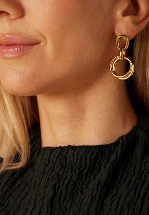 Boucles d'oreilles en or avec des anneaux circulaires entrelacés de différentes tailles. Surface lisse avec un fini brillant, portées avec un haut noir texturé.