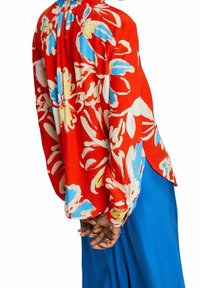 Blouse rouge à motif floral avec des accents bleus et jaunes ; coupe ample, manches longues, encolure froncée, associée à une jupe bleu uni.