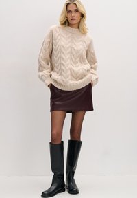 Beżowy sweter z grubym splotem i wysokim kołnierzem, bordowa skórzana mini spódnica oraz czarne kozaki za kolano. Gładkie tekstury i kontrastujące kolory.