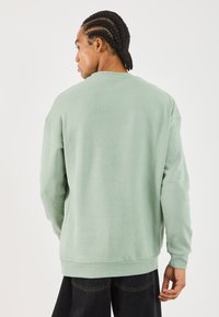 Hombre con trenzas que lleva una sudadera de manga larga verde claro y pantalones oscuros, de espaldas contra un fondo blanco liso.