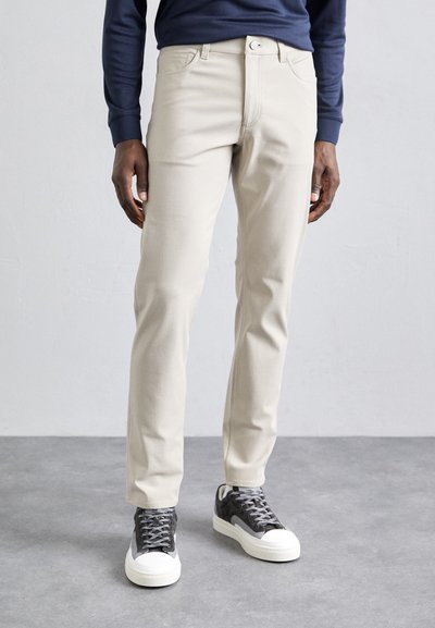 Pantalons en coton beige avec une coupe slim, poches avant et fermeture éclair, associés à des baskets grises avec des semelles blanches et des lacets à motifs.