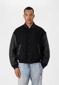 Veste universitaire noire avec manches en cuir noir, dotée d'un corps en laine texturé, d'un col côtelé et d'une fermeture à boutons. Logo notable sur la poitrine.