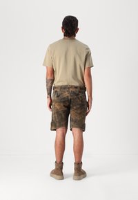 Beige T-Shirt mit kurzen Ärmeln, olivgrüne Shorts mit einer abgenutzten braunen Textur und braune Knöchelstiefel mit einer hellen Sohle.