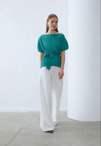 Top teal plissettato con maniche corte e vita legata, abbinato a pantaloni larghi bianchi e scarpe nere, su un pavimento liscio grigio chiaro.