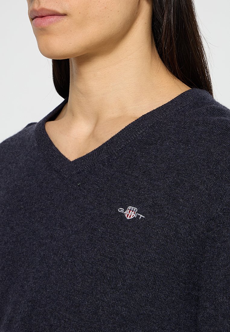 Navyblauer V-Ausschnitt-Pullover aus weichem Material mit einem kleinen gestickten Logo auf der Brust. Detaillierte Textur sichtbar.