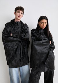 To modeller har på seg overdimensjonerte svarte satengponchoer med videre ermer og høye krager. Ponchoene har en teksturert overflate, kombinert med lyse denim-bukser.
