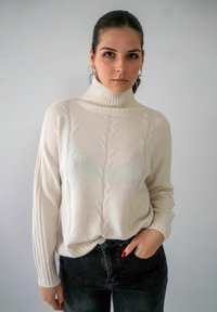 Crème gebreid sweater met een hoge hals, voorzien van een kabelpatroon en geribbelde mouwen. Gecombineerd met donkere jeans voor contrast.