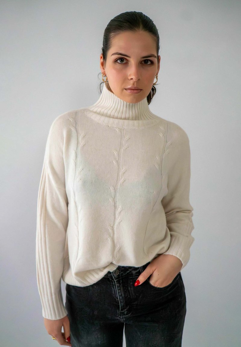 Crème gebreid sweater met een hoge hals, voorzien van een kabelpatroon en geribbelde mouwen. Gecombineerd met donkere jeans voor contrast.