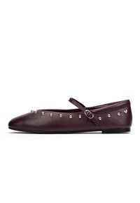 Scarpa piatta in pelle marrone scuro con borchie argentate lungo il bordo e una strap Mary Jane regolabile sopra il collo del piede.