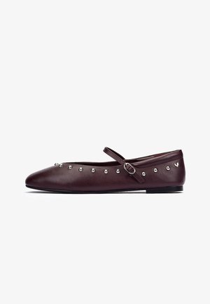 Zapato plano de cuero marrón oscuro con tachuelas plateadas a lo largo del borde y una correa ajustable de tipo Mary Jane en el empeine.