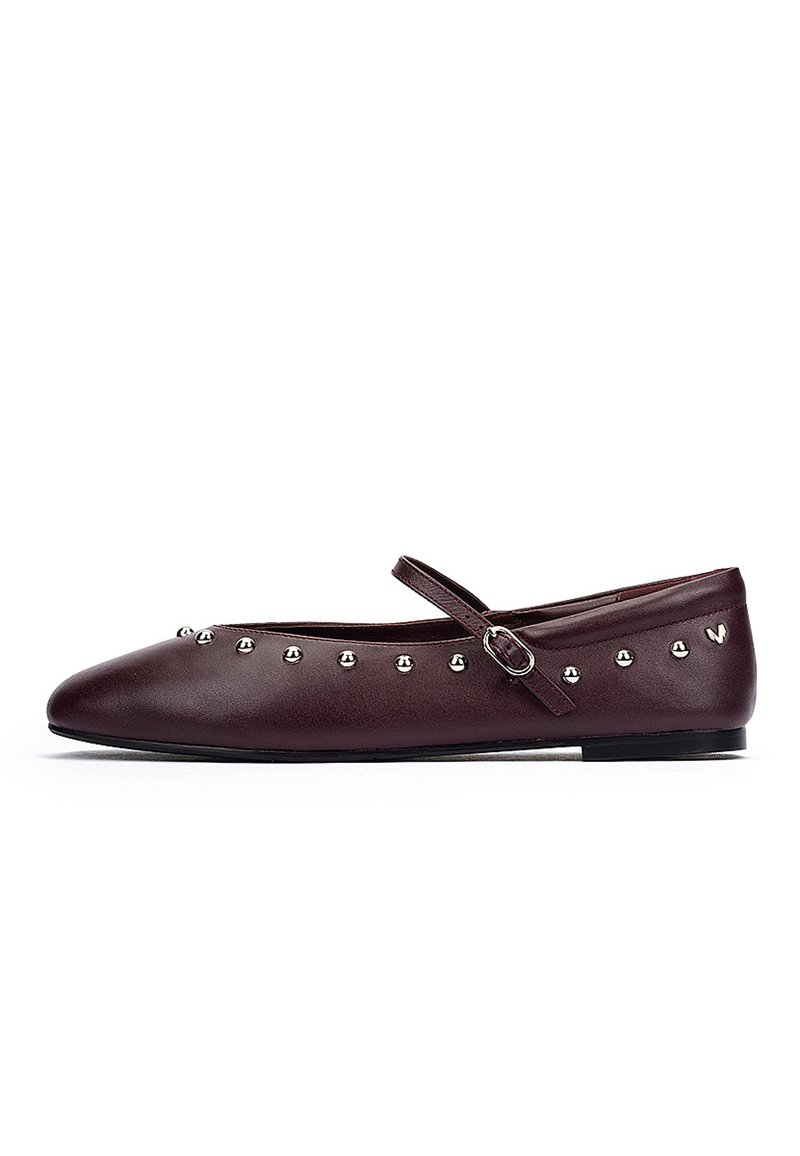 Scarpa piatta in pelle marrone scuro con borchie argentate lungo il bordo e una strap Mary Jane regolabile sopra il collo del piede.