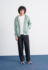 Um hoodie verde claro com fecho éclair por cima de uma camisa branca, emparelhado com calças de fato pretas com riscas laterais e sapatos esportivos cinzentos.