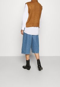 Pull sans manches en tricot marron porté sur une chemise blanche, associé à un pantalon culotte en denim bleu et des bottines noires. Tissus lisses et coupe décontractée.