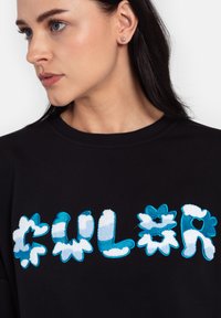 Czarna bluza z textured niebieskimi i białymi haftami kwiatowymi, z zabawnym, nieregularnym fontem wypisującym "CULUR". Zbliżenie na profil modela.
