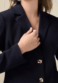 Blazer bleu marine avec une coupe ajustée, un col à revers cranté et des boutons dorés. Texture lisse, gros plan sur une main ajustant le détail du col.