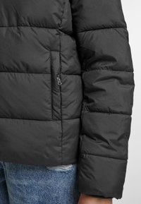 Svart puffjakke med quiltede seksjoner, med en glidelåslomme og en jevn tekstur. Brukt over slitte jeans.