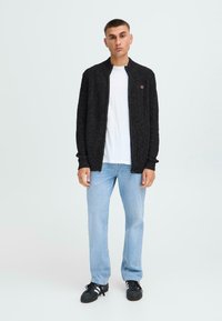 Cardigan nero lavorato a maglia con colletto alto, indossato sopra una t-shirt bianca. Abbinato a jeans azzurri chiari e sneakers nere. Dettaglio del logo discreto visibile.