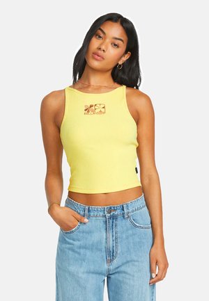 Frau trägt ärmelloses gelbes Crop-Top mit floralem Logo und hoch taillierte hellblaue Jeans, posiert mit einer Hand in der Tasche.