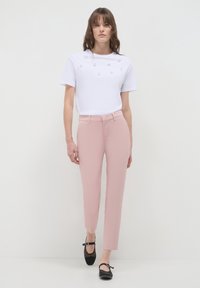 Woman wearing a white T-shirt with pearl embellishments, pink tailored pants, and black Mary Jane flats walking against a plain background.