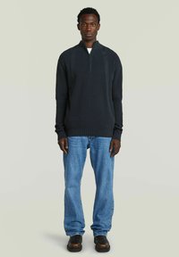 Jonge man met kort krullend haar, gekleed in een zwarte sweater met rits tot de helft, lichtblauwe jeans en zwarte schoenen, staande tegen een lichtgrijze achtergrond.