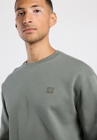 Grijze sweatshirt met ronde hals, ribbels bij de mouwen en een klein, getextureerd logo op de linkerkant van de borst. Zacht katoenmateriaal, reguliere pasvorm.
