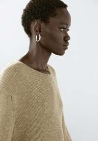 Maglione beige lavorato a maglia con scollo ampio, caratterizzato da una texture a coste. Indossato con grandi orecchini a cerchio in argento. Pelle liscia e capelli corti visibili.