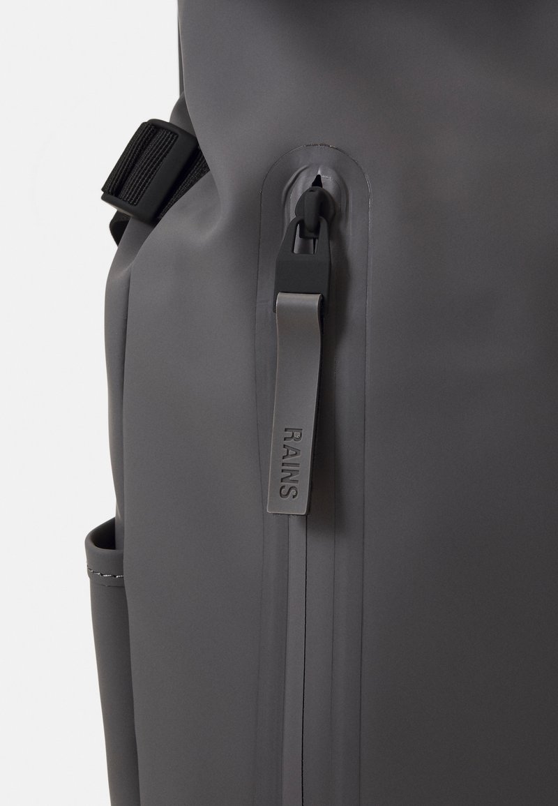 Mochila impermeable gris con una superficie lisa, un bolsillo lateral y un tirador de cremallera de la marca con "RAINS" grabado.